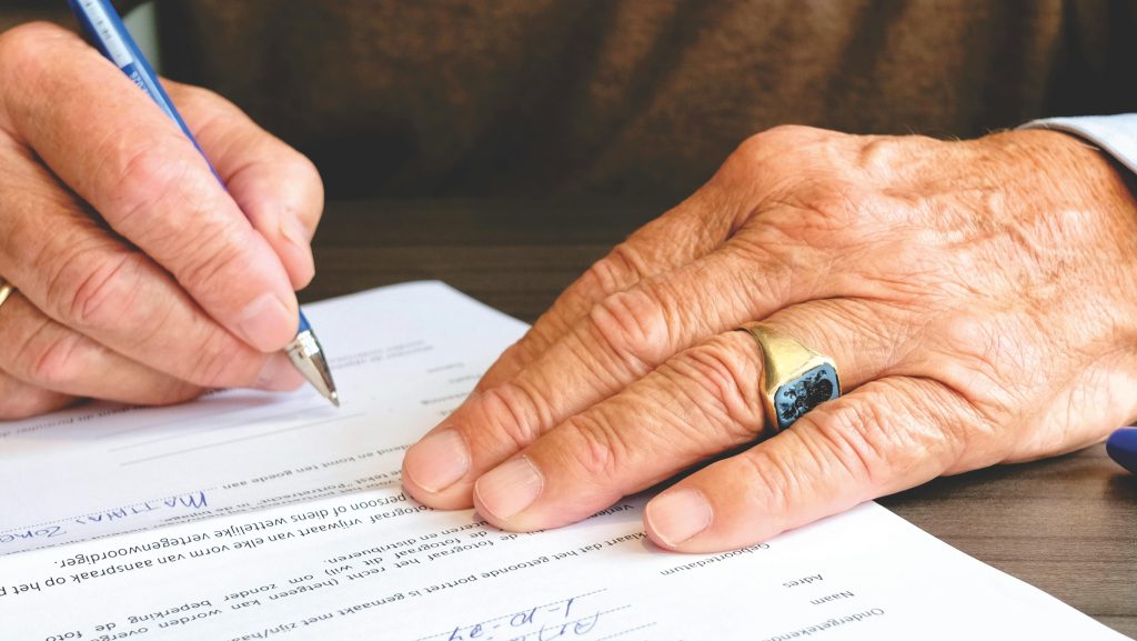 Layanan Pertanahan & Properti Close-up of a senior adult signing a legal document with a focus on hand and gold ring.